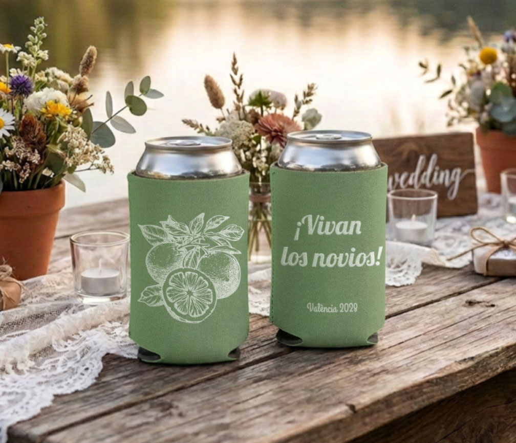 Close-up of sage green personalized wedding can coolers on silver cans, citrus line art and ¡Vivan los novios!, collapsible neoprene wedding favors on a wooden reception table.