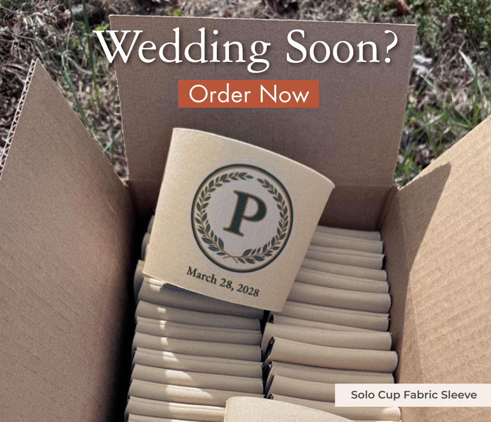 Open box of kraft solo cup fabric sleeves stacked flat, one sleeve upright with a green laurel monogram P and March 28, 2026, a personalized wedding favor ready to ship.